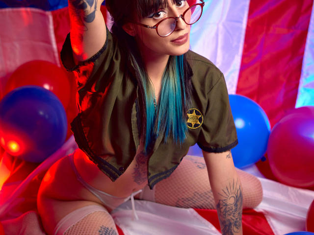 TatsumiLee with blue-tipped hair, glasses, green jacket, white panties, fishnet stockings, tattoos, kneeling, reaching up, red and blue balloons, red and white background, sexy, confident.