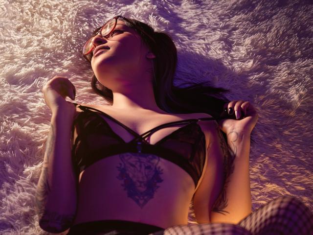 TatsumiLee lying on a fluffy white rug, wearing black strappy lingerie, showing a chest tattoo above her tits, holding her hair, looking up with a relaxed expression.