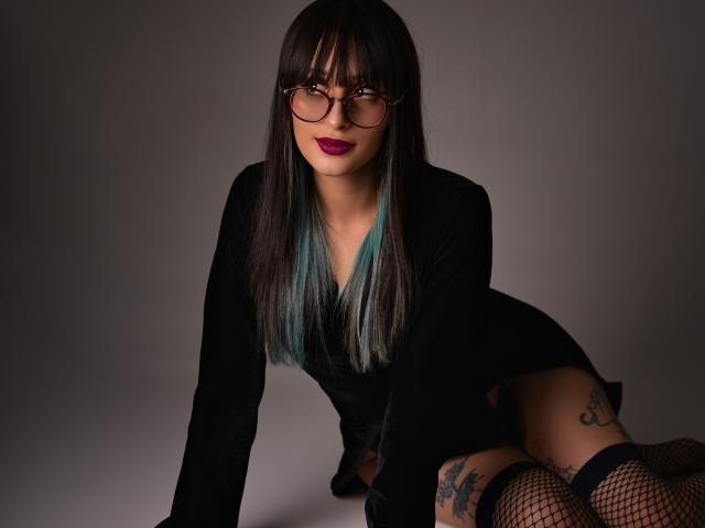 TatsumiLee, con capelli scuri, occhiali e rossetto viola, indossa un abito nero e calze a rete, è piegata in avanti, mostrando dei tatuaggi sulla coscia, con le punte dei capelli colorate di blu..