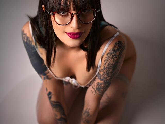 TatsumiLee mit Brille, dunklem Lippenstift und Tätowierungen, kniend, die Brüste zeigen, in Dessous, mit einem leichten Lächeln nach oben schauend. Ihre Arme und Oberschenkel sind mit aufwendigen Tätowierungen bedeckt..