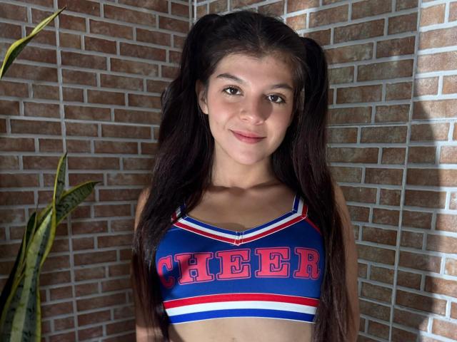 RilleyFoyx with long dark hair in pigtails, wearing a blue and red cheerleader top, smiling at the camera, showing her small tits. Brown brick wall in the background.