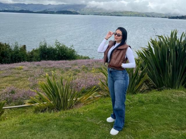 IsabellaJames stands with hand on forehead, wearing a brown vest, white shirt, blue jeans, and white sneakers, by a lake with purple flowers and green plants.