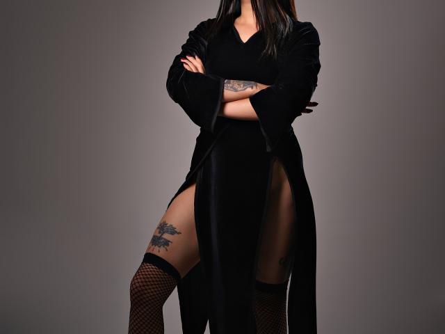 TatsumiLee è in piedi, con le braccia incrociate, indossa un abito nero con una profonda spaccatura, calze a rete nere e ha dei tatuaggi sulla coscia destra e sul braccio sinistro. I suoi capelli neri sono sciolti..