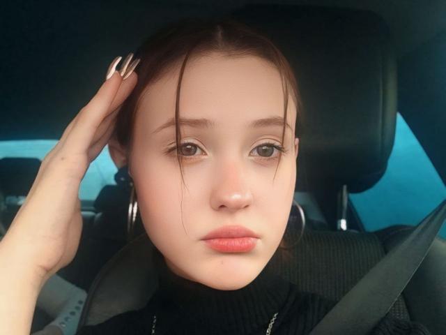 LillyDupass, met een lichte huid, bruine ogen en roze lippen, draagt een zwarte coltrui en zit in een auto. Ze heeft haar hand in haar haar, lange nagels, oorringen en draagt een veiligheidsgordel..