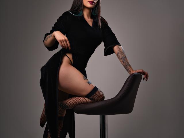 TatsumiLee mit Tattoos, trägt ein schwarzes Kleid, Fischernetzstrümpfe und sitzt auf einem dunklen Stuhl, wobei ihre Oberschenkel und Vulva sichtbar sind. Ein Bein ist auf dem Stuhl, sie trägt dunklen Lippenstift und posiert verführerisch..