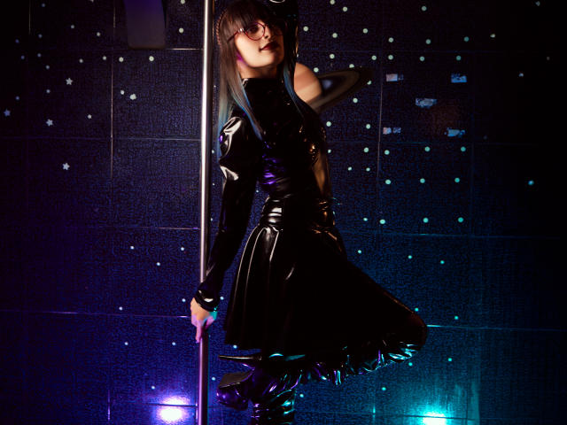 TatsumiLee trägt ein glänzendes, schwarzes Latexdress, hält mit einer Hand einen Stab, trägt schwarze Handschuhe und Stiefel, hat dunkles Haar, blaues Licht, steht und nimmt eine verführerische Pose ein..