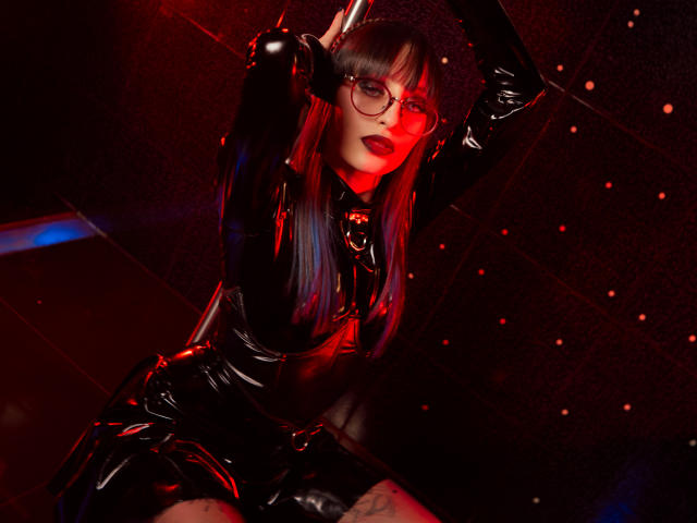 TatsumiLee mit langem, schwarzem und blauem Haar, trägt ein glänzendes schwarzes Latexdress und eine Brille, die Arme sind erhoben, sie sieht verführerisch aus, rote und schwarze Beleuchtung, ein Tattoo ist auf dem Oberschenkel sichtbar..