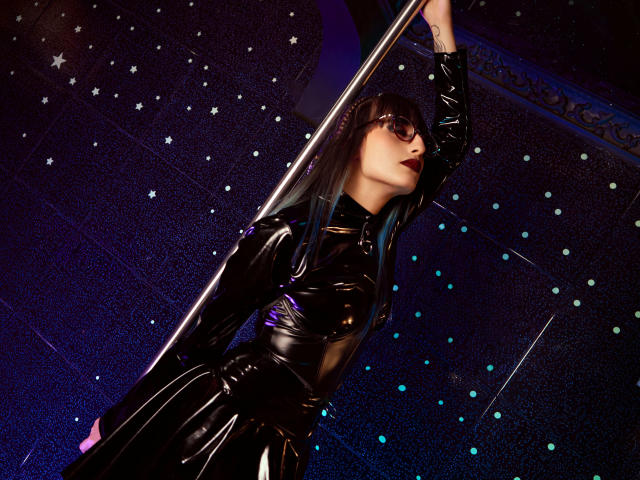 TatsumiLee con un abito di lattice nero, che tiene un palo, indossa occhiali da sole scuri, rossetto rosso, lunghi capelli neri, braccio alzato, che guarda in alto, materiale lucido, illuminazione viola e blu..