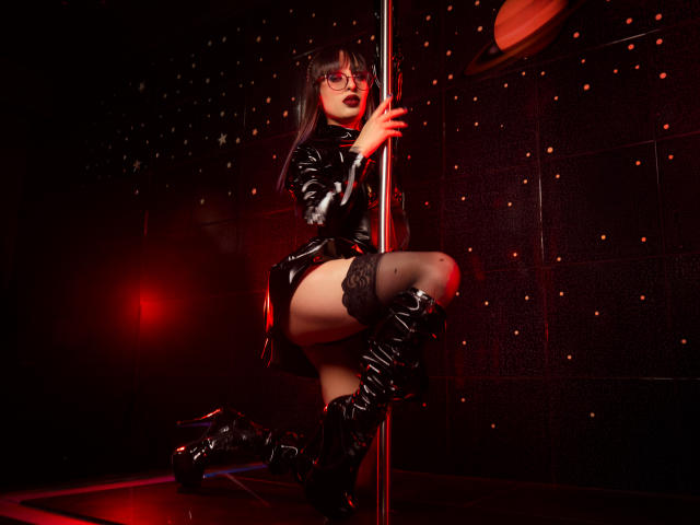 TatsumiLee in einem schwarzen Latexrkleid, kniehohen Stiefeln, Spitzenstrümpfen, einer Brille und rotem Lippenstift, kniet vor einem Pfosten, zeigt ihren Hintern, mit einer Hand auf dem Pfosten, in einem rot beleuchteten Raum..