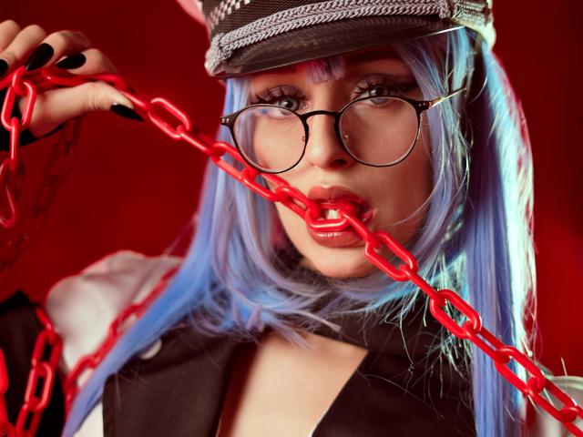 TatsumiLee mit blauen Haaren, einer Brille und einem Hut, beißt an einer roten Kette, wobei ihre vollen, roten Lippen und schwarzen Nägel auffallen. Ihre Brüste sind teilweise sichtbar..