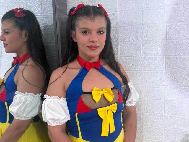 RilleyFoyx has long brown hair in pigtails with red bows, wearing a blue, yellow, and red costume with cutout boobs, white puffy sleeves, and a yellow bow.