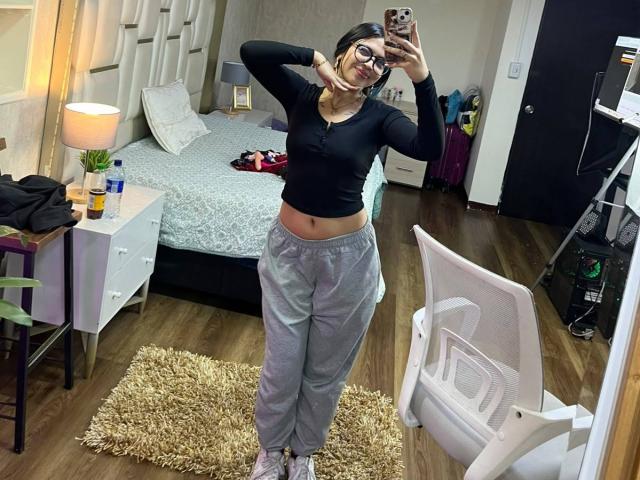 NyraVanel, usando óculos, com uma blusa de manga comprida preta, calças de moletom cinzas e tênis rosa, está em pé em um quarto, com os braços atrás da cabeça, tirando uma selfie no espelho, mostrando a parte do abdômen e o bumbum..