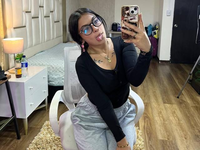 NyraVanel con capelli neri, occhiali, top nero a maniche lunghe, pantaloni grigi, lingua fuori, che si fa un selfie allo specchio, tatuaggi sul petto, sedia bianca, pavimento di legno, telefono con custodia decorativa..