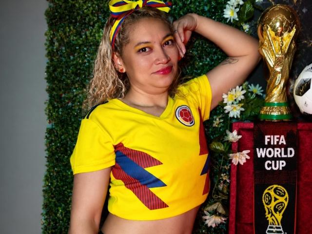 MeganLatin, com cabelo loiro cacheado, vestindo uma camisa de futebol amarela e azul, mostrando seus seios, com o braço levantado, em pé ao lado de um troféu da Copa do Mundo da FIFA..