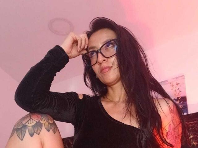 ZarayFeet hat langes, schwarzes Haar, eine Brille und ein schwarzes Samintop mit ausgestanzten Ärmeln. Man sieht ihr ein tätowiertes Knie und eine Halskette. Sie blickt leicht nach oben und hat ein schmollendes Gesicht..