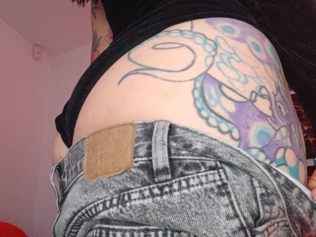 ZarayFeet's butt with tattooed side, showing a colorful octopus design, in worn-out blue jeans, black top, and a tan leather belt patch, in a dimly lit room.