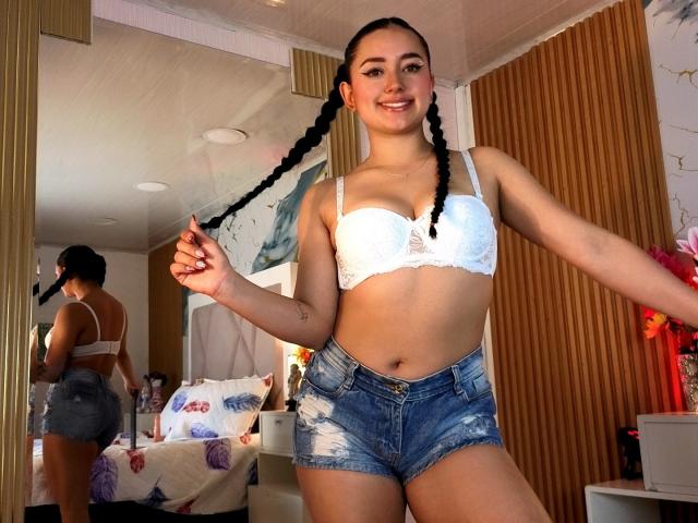 MiiaRousse con i capelli intrecciati, un reggiseno di pizzo bianco e pantaloncini di jeans, che tiene una treccia, sorride, mostrando i suoi seni, in una camera da letto con uno specchio che riflette il suo sedere..