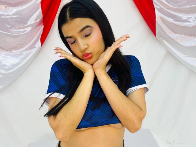 Iris Blair mit dunklem Haar, einem blauen Crop-Top, den Händen am Kinn, geschürzten roten Lippen, rosa Nägeln, die nach unten schaut, mit einem weiß-roten Hintergrund. Ihre Brüste sind vom Top bedeckt..