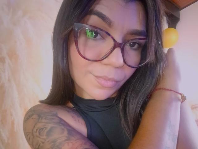 ValeryPierce with dark brown hair, glasses, black top, and tattooed arm, holding a yellow lemon, smiling, green eye reflection, red bracelet, and subtle makeup.