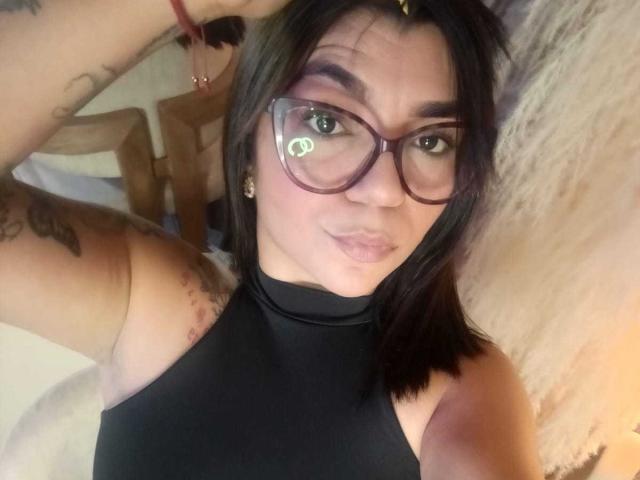 Valery Pierce con gli occhiali, maglione nero a collo alto, labbra piene, tatuaggi sulle braccia, capelli castano scuro, pelle tendente al rosa, che guarda la fotocamera, braccio destro alzato, braccialetto rosso al polso..