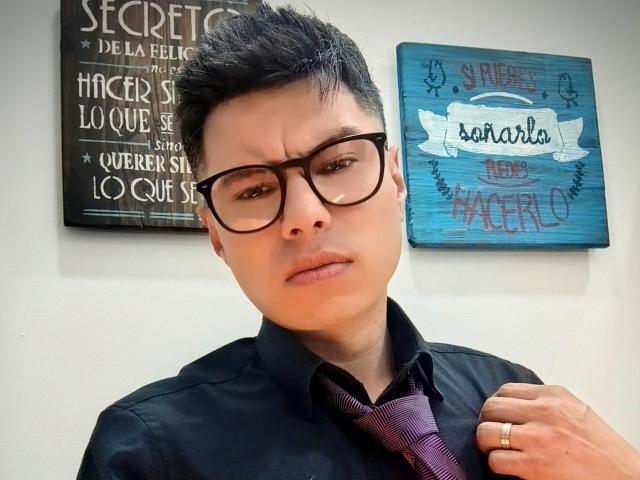 AquilesTaylor with black-framed glasses, short spiked hair, black shirt, purple tie, serious expression, slightly furrowed brows, right hand holding tie, left hand with gold ring.