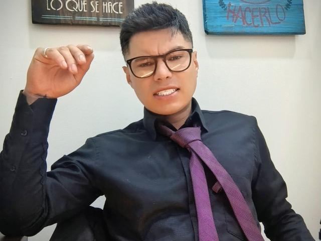 AquilesTaylor met kort zwart haar, een bril, een zwart shirt, een paarse stropdas en een opgetrokken vuist, met een licht geïrriteerde uitdrukking en samengeknepen tanden..