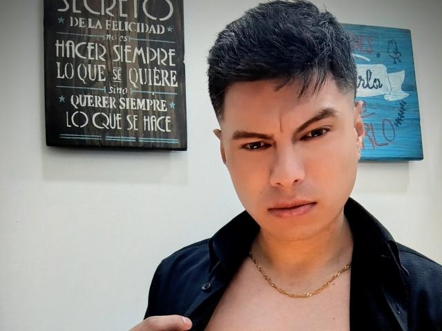 Aquiles Taylor, avec ses cheveux noirs courts, une chaîne en or et une chemise noire, regarde vers le bas avec un léger froncement de sourcils. On peut voir son teint mat et ses sourcils bien dessinés..