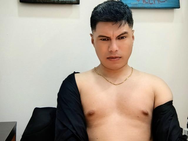 AquilesTaylor è un uomo asiatico senza camicia, con capelli neri corti, una catena d'oro e un'espressione seria, che indossa una giacca nera portata sulle spalle, lasciando scoperte le sue piccole e nude mammelle..