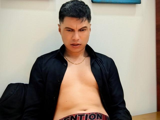 Aquiles Taylor, con i capelli neri e corti, guarda intensamente, con la camicia aperta, che rivela un petto liscio, abbronzato e nudo, e una cintura rossa con la scritta "GATEF". Indossa una collana d'oro..