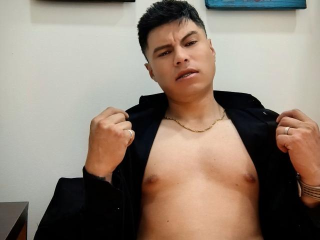 AquilesTaylor, shirtless, black jacket open, small chest, gold chain, short black hair, slightly angry expression, holding jacket edges, ring on right hand, tattoo on left wrist.