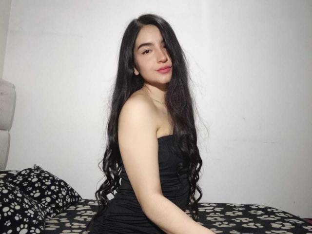 Jade Harpper, con lunghi capelli neri, pelle chiara e un sorriso carino, indossa un abito nero senza spalline, seduta su un letto con lenzuola a motivi bianchi e neri..