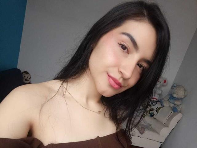 JadeHarpper has long black hair, brown eyes, and pink lipstick. She's smiling with a slight tilt, wearing an off-shoulder brown top, showing her bare shoulders and small tits.