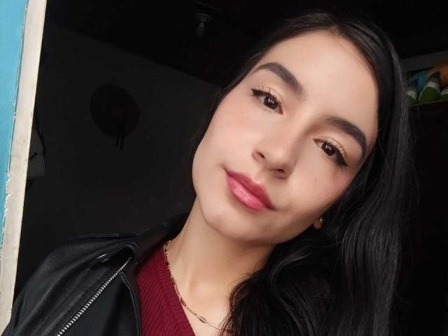 Jade Harpper ha lunghi capelli neri, occhi marroni e rossetto rosa. Indossa una giacca di pelle nera sopra un maglione rosso, con una sottile collana dorata. Sorride dolcemente..