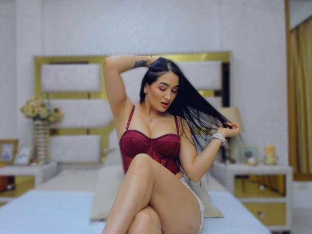 PamelaLiss, avec de longs cheveux noirs, un soutien-gorge rouge et des sous-vêtements blancs, assise sur un lit, touchant ses cheveux, un tatouage sur le bras, sexy, pulpeuse, avec de grandes seins, des hanches moyennes, dans une chambre, détendue..