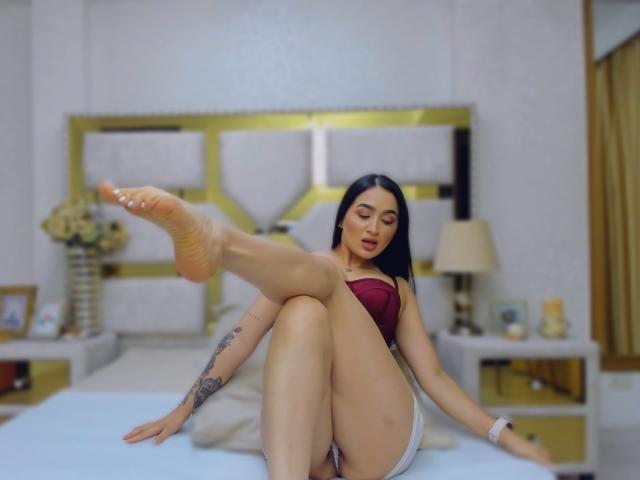 PamelaLiss mit langen, schwarzen Haaren, einem roten, trägerlosen Top, weißen Shorts, einem Tattoo am rechten Arm, mit angehobenem Bein, wobei ihr nackter Fuß zu sehen ist, sitzend auf einem Bett, nach unten schauend, mit rotem Lippenstift..