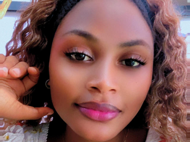 LizzyClem has curly brown hair, pink lipstick, and long eyelashes, smiling with a hand near her ear. Her dark brown eyes and smooth skin are visible.