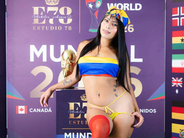 LeiaLaurent, com longos cabelos pretos, vestindo um top tipo bandeau amarelo, azul e vermelho, uma tanga amarela e meias vermelhas até a coxa, segurando um troféu dourado, exibindo seus seios e nádegas..