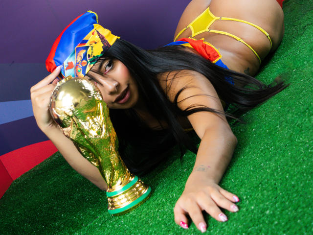 LeiaLaurent, avec de longs cheveux noirs, portant un string jaune et un chapeau bleu-rouge-jaune, allongée sur de l'herbe verte, tenant un trophée FIFA doré, les fesses en l'air, regardant l'appareil photo..