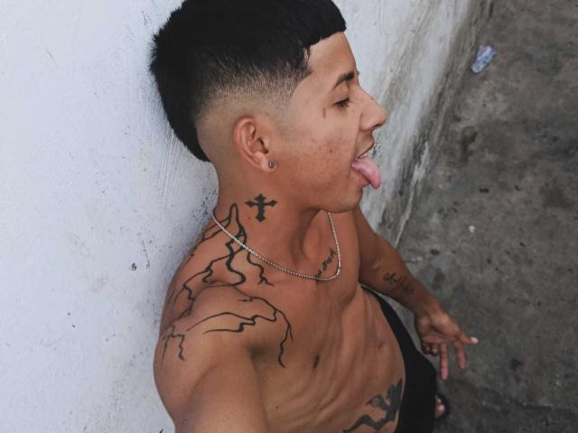 JuliianWolf with short black hair, tongue out, shirtless, tattoos on neck and shoulder, chain necklace, earrings, and a cross tattoo on the left side of his neck.