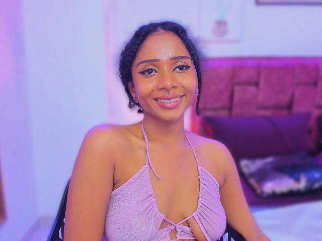 Megan Williams, con i capelli neri ricci, la pelle marrone, indossa un top rosa a halter, sorride con un piercing al naso, mostrando il suo seno piccolo e il suo viso carino. Illuminazione viola e rosa..