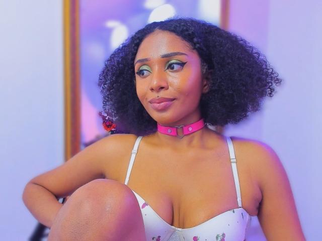 MeganWilliams with curly black hair, wearing a pink choker, white tank top with pink hearts, and green eye makeup, sitting with knee up, showing her boobs.