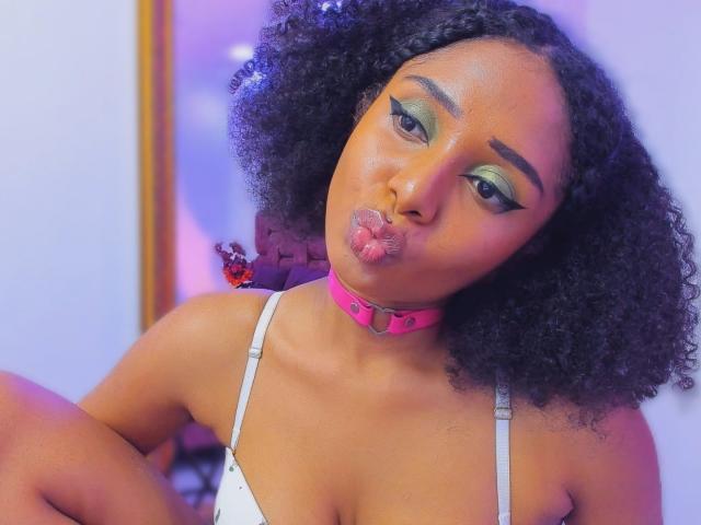 MeganWilliams with curly black hair, pink choker, white tank top, pouty lips, green eyeshadow, and bold eyeliner. Boobs are partially visible. She's making a kissy face.