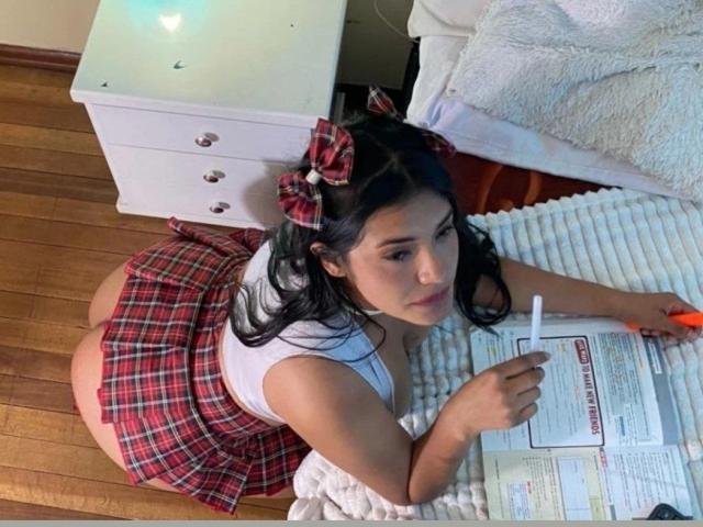 ArianithaXox with dark hair, red plaid skirt, white top, and red bow, lying on her stomach, writing with an orange pen, showing her big ass, on a wooden floor.