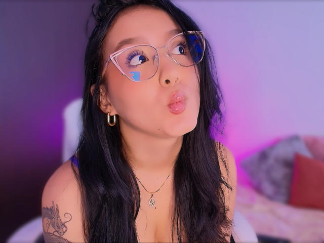 SophiaBrowm with pouty lips, wearing clear glasses, black hair, hoop earrings, necklace, tattoo on right shoulder, blue face paint, and a blue eye mask, in a bedroom.