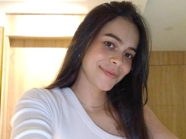 Alana Blake, avec ses longs cheveux foncés, sa chemise blanche, son sourire discret et son petit collier, présente un visage adorable et une expression douce, avec un éclairage subtil..