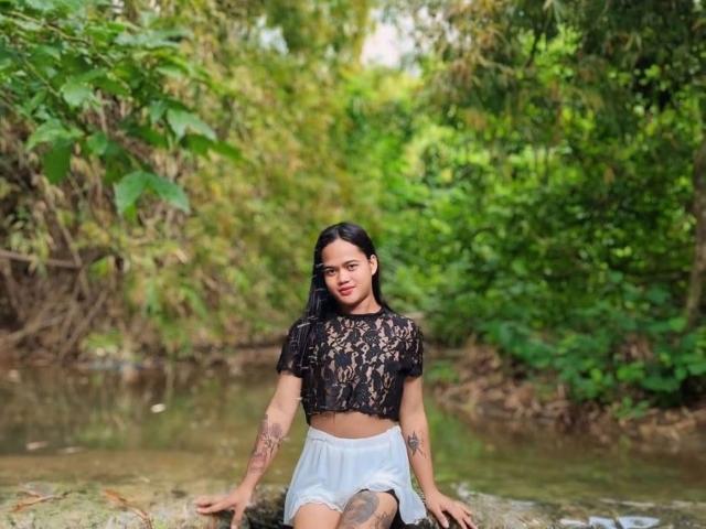 Una mujer con tatuajes, una camiseta de encaje negro y pantalones cortos blancos, sentada sobre una roca junto a un arroyo, con un fondo de bosque verde, cabello negro largo, lápiz labial rojo, mirando a la cámara..