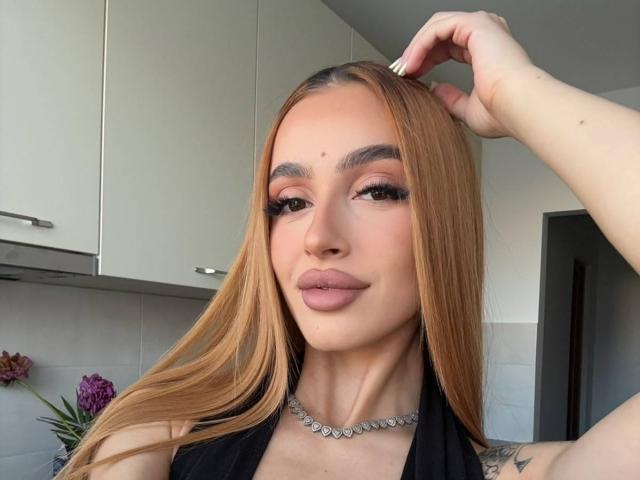 MarissaFex, avec ses longs cheveux blonds, ses lèvres roses et pulpeuses, et son collier de diamants, lève la main à sa tête, exhibant sa poitrine généreuse et un tatouage sur son bras..