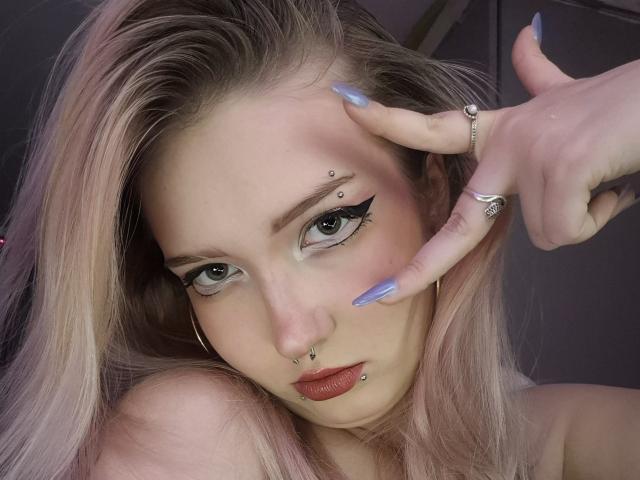 PalmiraEsquinaldo, com cabelo rosa, olhos verdes, delineador preto, piercing no nariz, piercing no lábio e unhas azuis, fazendo um sinal de paz com a mão adornada com anéis. Seios parcialmente visíveis..