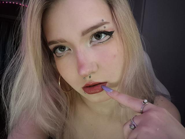 PalmiraEsquinaldo, com cabelo loiro, piercings no nariz e lábio superior, batom roxo, dedo na boca, unhas roxas e brincos de argola, olhando para a câmera..