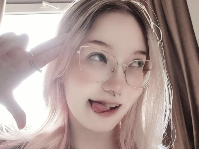 PalmiraEsquinaldo, com cabelo rosa pálido, óculos, piercing no nariz, piercing no septo e a língua de fora, fazendo um sinal de paz com um dedo com anel. Suas nádegas não estão visíveis..
